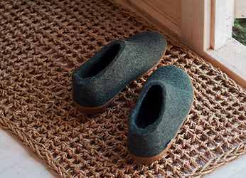 A pair of glerups shoes in the color Forest green placed on a woven doormat near a wooden doorway. The shoes have a natural rubber sole.