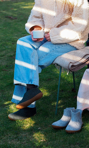 A person sitting outdoors on a chair with a blanket draped over it, holding a cup. The person is wearing glerups shoes in the color Charcoal grey, and an additional pair of glerups boots in the color Grey is placed on the grass nearby.
