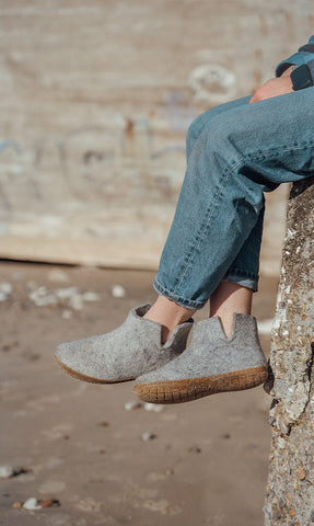 A person sitting on a sandy surface wearing glerups boots in the color Grey with natural rubber sole. The background shows a textured rock or wall.