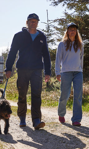 Two persons walking on a forest path, each wearing glerups shoes. One is holding a dog’s leash, and trees and sunlight are visible in the background.