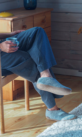 A person sitting indoors on a wooden chair, holding a cup and wearing Grey glerups shoes. The setting includes wooden furniture and soft natural light.