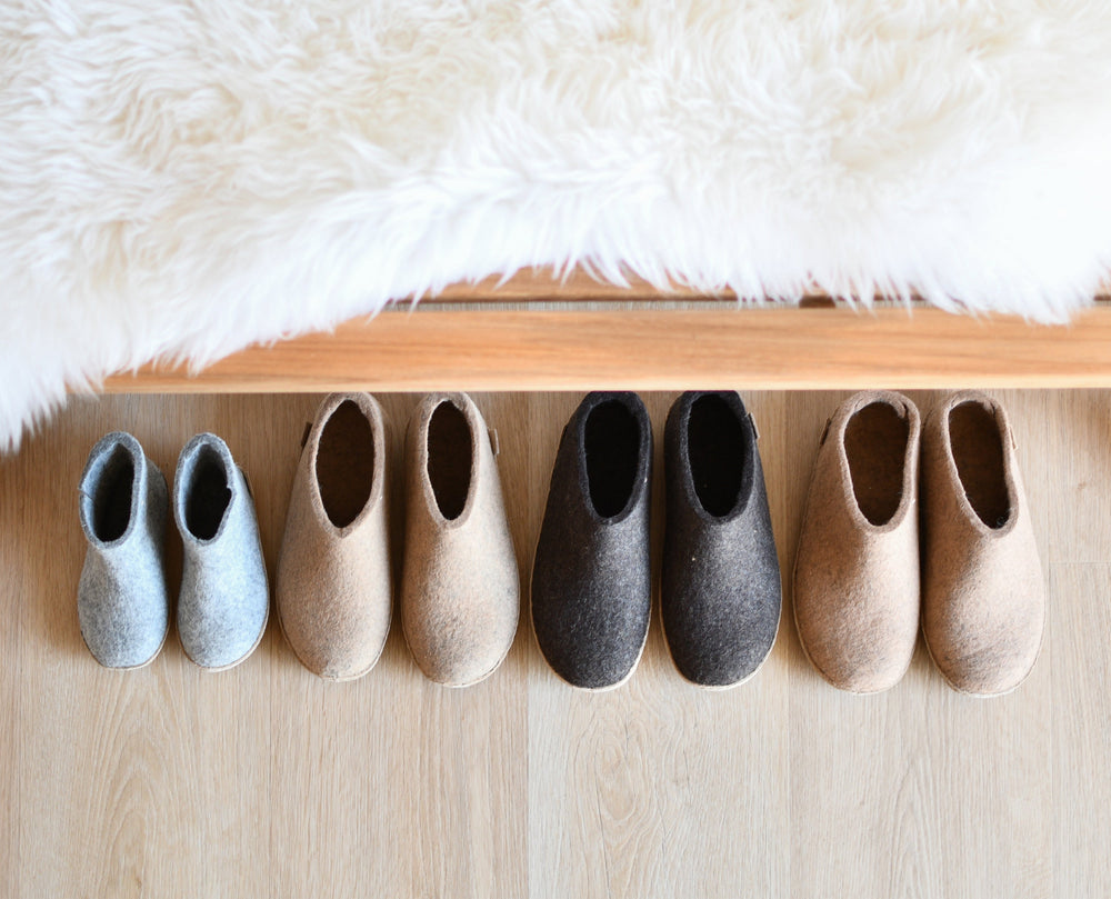 Several pairs of glerups shoes in different neutral colors arranged in a row on a light wooden floor. A fluffy white blanket hangs over the edge of a wooden bed frame above the shoes.