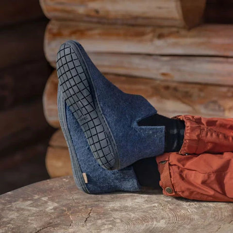 A person is wearing glerups boots in the color Denim blue with black rubber soles, with feet resting on a wooden surface.