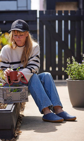 A person with blond hair and a cap is sitting outdoors on a patio holding a small plant pot, wearing casual clothing and glerups slip‑os in the color Denim blue. Plants and gardening items are placed around the person.