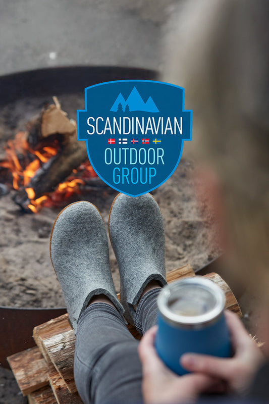 Several individuals are sitting in tall grass on outdoor chairs with their legs extended, each wearing glerups shoes in different colors. A large blue badge graphic in the center reads ‘SCANDINAVIAN OUTDOOR GROUP’ with small Nordic flags displayed above the text.