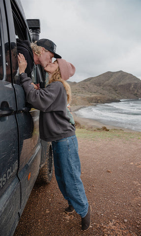 Two persons standing beside a vehicle near a coastal landscape, leaning toward each other and kissing. One individual is wearing glerups boots in the color Chestnut brown while standing on a dirt road with mountains and ocean in the background.